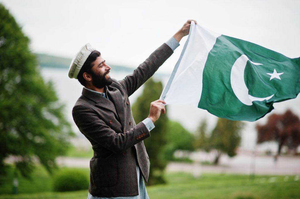Beard pakistani man wear pakol hat and jacket hold Pakistan flag.