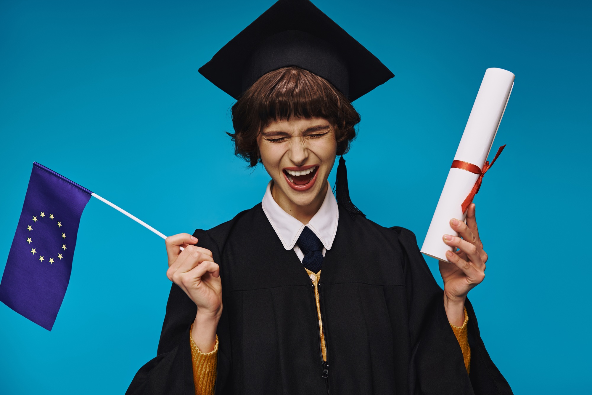 excited graduated college girl in gown and academic cap with diploma and EU flag on blue backdrop